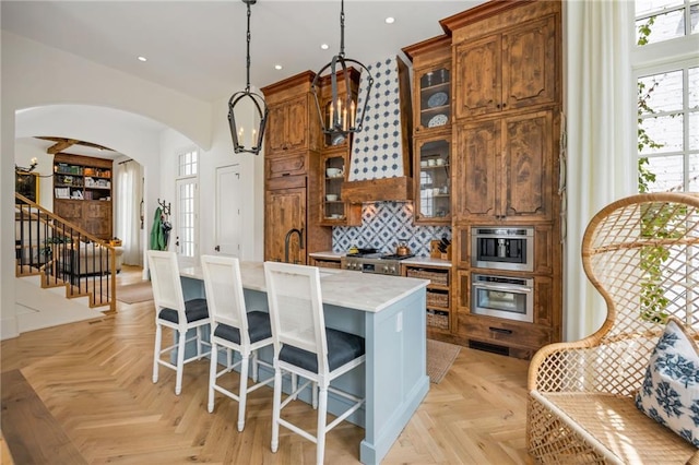 11277 Serenbe Ln, Chattahoochee Hills, GA 30268 - kitchen featuring arched walkways, an island with sink, brown cabinets, decorative light fixtures, and decorative backsplash