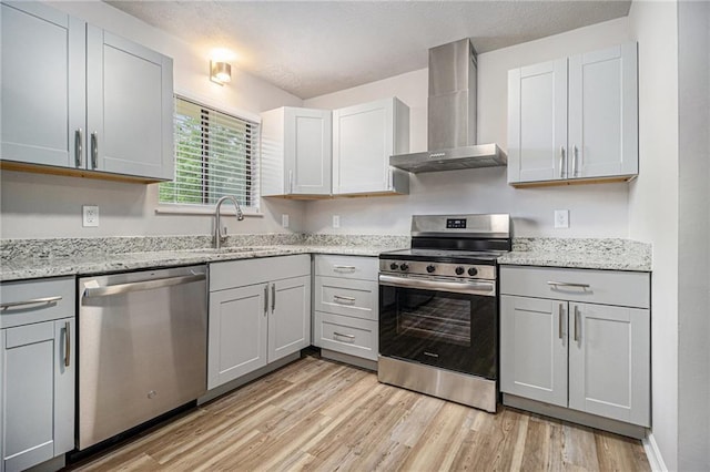 kitchen featuring wall chimney range hood, appliances with stainless steel finishes, light stone countertops, a sink, and light wood finished floors