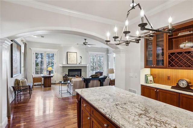 kitchen featuring brown cabinetry, light stone countertops, dark wood finished floors, ceiling fan with notable chandelier, and arched walkways