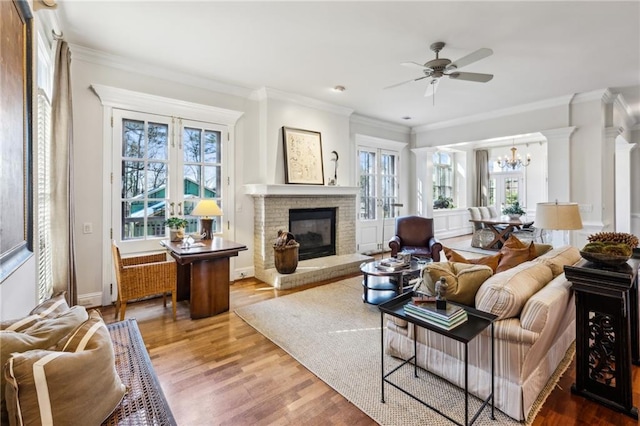 living area with ornamental molding, ceiling fan with notable chandelier, wood finished floors, baseboards, and a brick fireplace