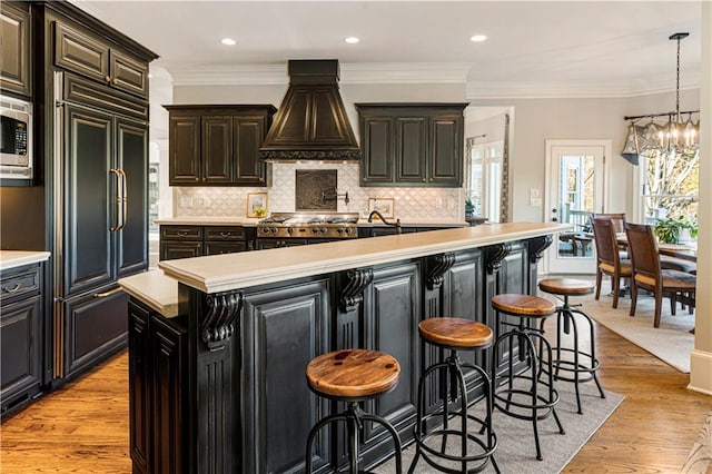 kitchen with a kitchen breakfast bar, an island with sink, decorative light fixtures, paneled built in fridge, and custom range hood