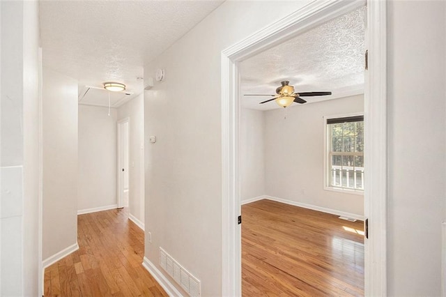 corridor with a textured ceiling, wood finished floors, visible vents, baseboards, and attic access