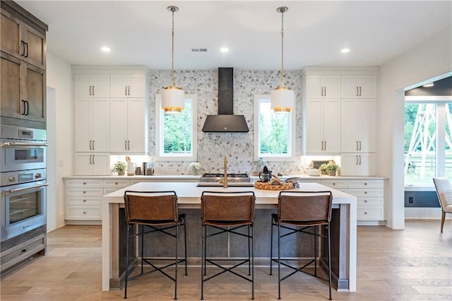 kitchen with a kitchen bar, light countertops, double oven, wall chimney exhaust hood, and an island with sink