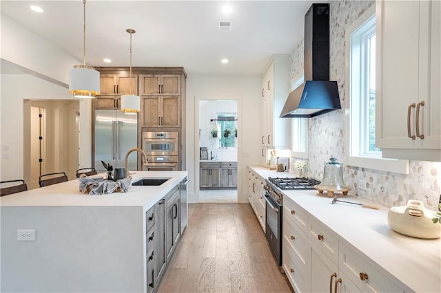 kitchen featuring wall chimney range hood, high quality appliances, light countertops, a sink, and light wood finished floors