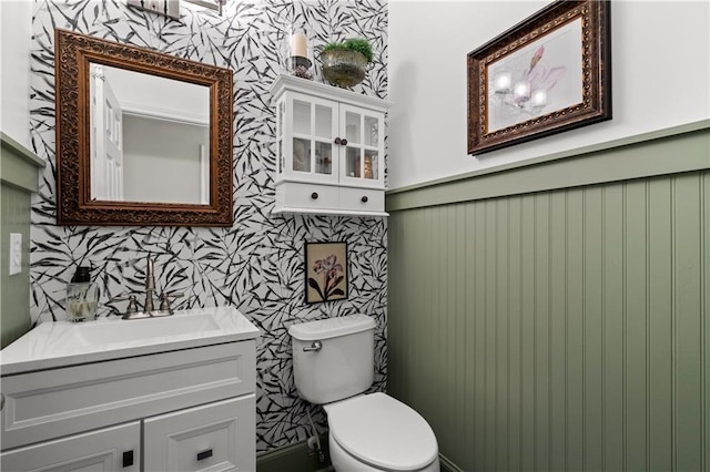 half bath featuring a wainscoted wall, toilet, vanity, and wallpapered walls