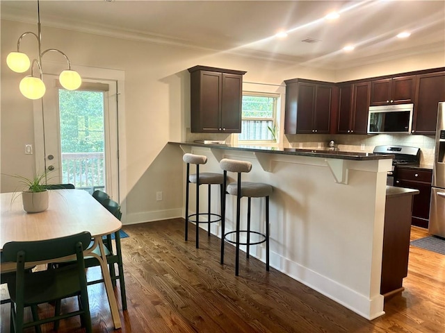 kitchen with appliances with stainless steel finishes, a breakfast bar area, dark countertops, backsplash, and dark wood finished floors