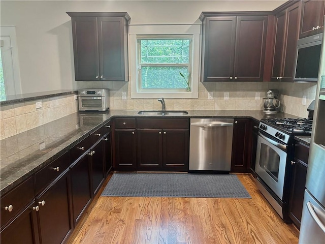 kitchen featuring appliances with stainless steel finishes, a sink, dark stone countertops, light wood-type flooring, and backsplash