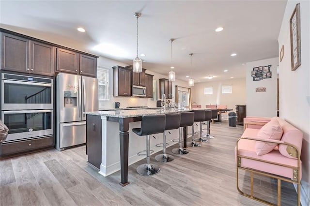 kitchen featuring appliances with stainless steel finishes, dark brown cabinets, light stone counters, an island with sink, and decorative light fixtures