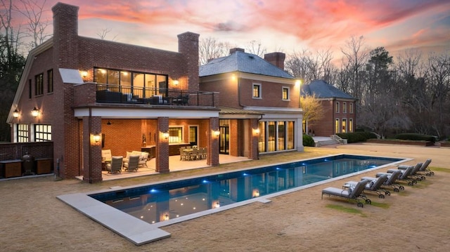 back of property at dusk with brick siding, a chimney, a balcony, a patio area, and an outdoor pool