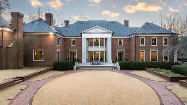 back of house at dusk with brick siding, a high end roof, a patio area, and a chimney