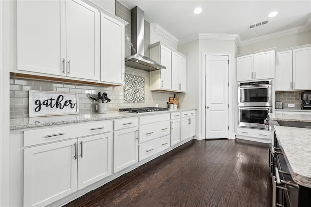 kitchen featuring appliances with stainless steel finishes, white cabinets, ornamental molding, light stone counters, and wall chimney exhaust hood