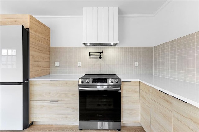 kitchen featuring wall chimney range hood, stainless steel electric range oven, light brown cabinetry, and freestanding refrigerator