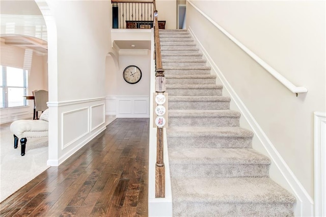 stairs with arched walkways, wood-type flooring, wainscoting, and a decorative wall