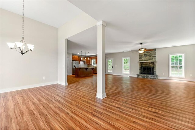 unfurnished living room with a ceiling fan, light wood-style flooring, a stone fireplace, a chandelier, and healthy amount of natural light