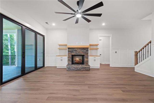 unfurnished living room featuring a ceiling fan, light wood-type flooring, stairway, a decorative wall, and a stone fireplace