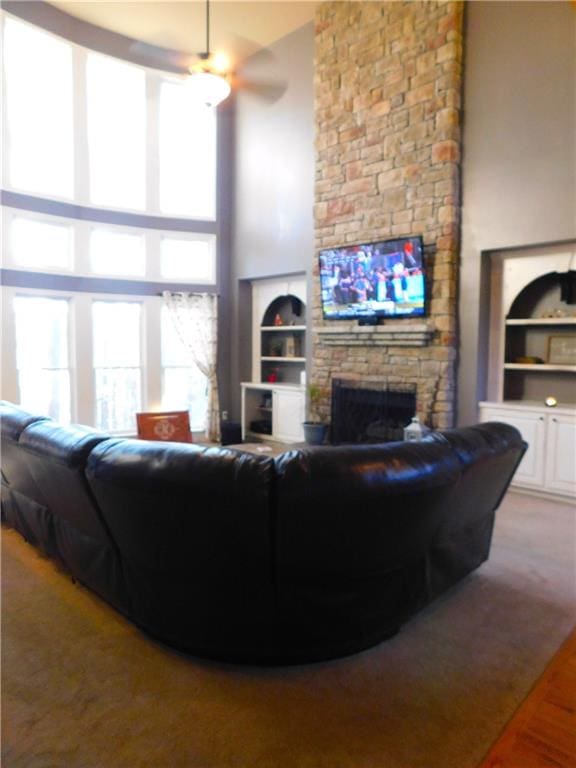 living room featuring a stone fireplace, ceiling fan, built in shelves, and a high ceiling