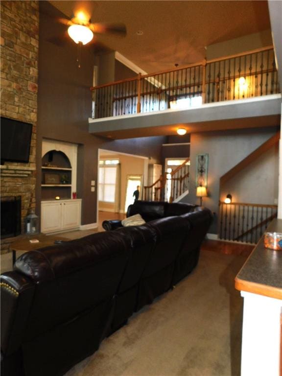 living room featuring carpet, a towering ceiling, built in shelves, ceiling fan, and a stone fireplace