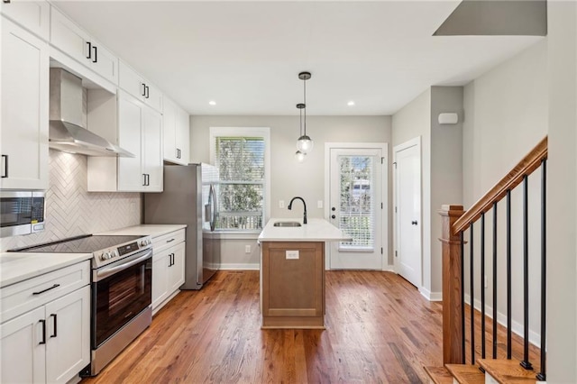 kitchen with stainless steel appliances, wall chimney exhaust hood, a kitchen island with sink, backsplash, and light wood finished floors