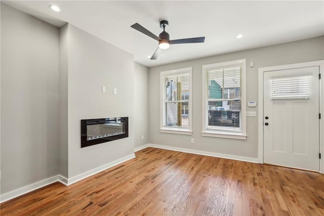 unfurnished living room featuring a glass covered fireplace, wood finished floors, baseboards, a ceiling fan, and recessed lighting