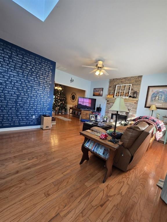 living room with wood-type flooring, a stone fireplace, ceiling fan, and a skylight