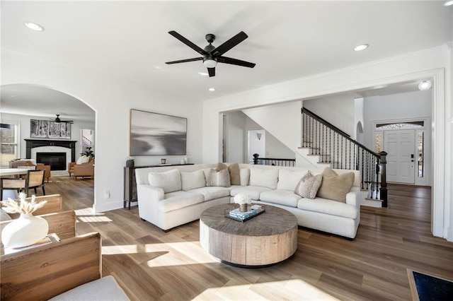 living room with arched walkways, a ceiling fan, a glass covered fireplace, stairs, and wood finished floors