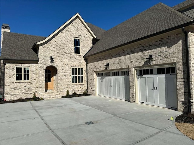 french country style house featuring brick siding, an attached garage, and roof with shingles