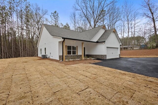 view of front of house with central AC unit, driveway, an attached garage, a shingled roof, and stone siding