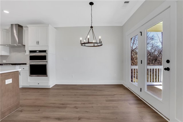 kitchen featuring wall chimney exhaust hood, light wood-style flooring, decorative light fixtures, stainless steel double oven, and white cabinetry