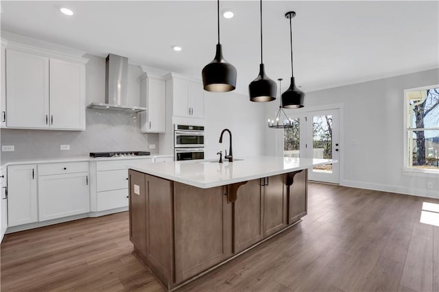 kitchen with white cabinets, wall chimney exhaust hood, a center island with sink, and light countertops