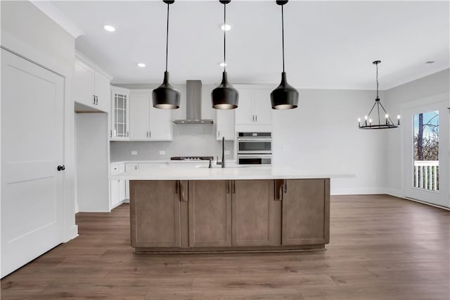 kitchen featuring a spacious island, white cabinetry, light countertops, wall chimney range hood, and glass insert cabinets