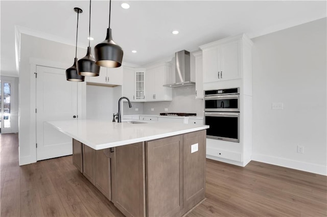 kitchen with light countertops, glass insert cabinets, white cabinets, a sink, and wall chimney exhaust hood