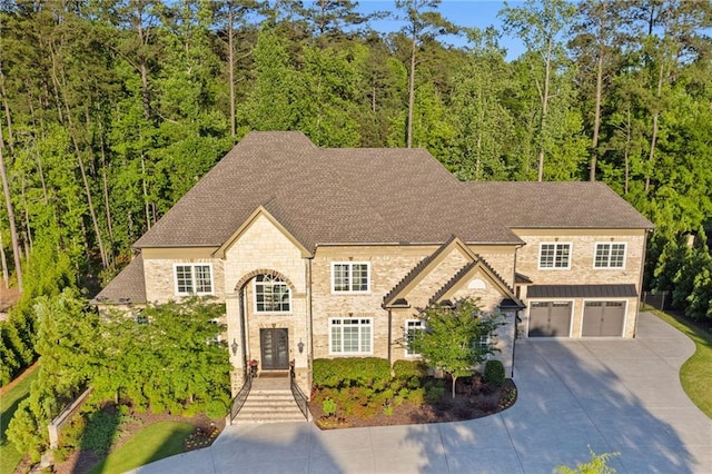view of front facade with an attached garage, stone siding, a wooded view, concrete driveway, and a shingled roof