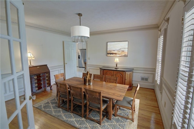 dining area with crown molding, light wood-style floors, french doors, a decorative wall, and a wainscoted wall