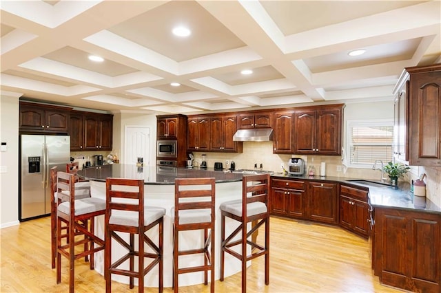 kitchen featuring appliances with stainless steel finishes, under cabinet range hood, a sink, a kitchen breakfast bar, and dark countertops