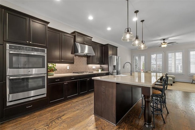 kitchen with a breakfast bar, stainless steel appliances, dark brown cabinetry, an island with sink, and decorative light fixtures