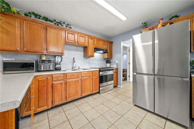 kitchen with light countertops, appliances with stainless steel finishes, brown cabinetry, a sink, and under cabinet range hood