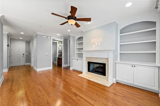 unfurnished living room with light wood-type flooring, built in shelves, a premium fireplace, ceiling fan, and crown molding