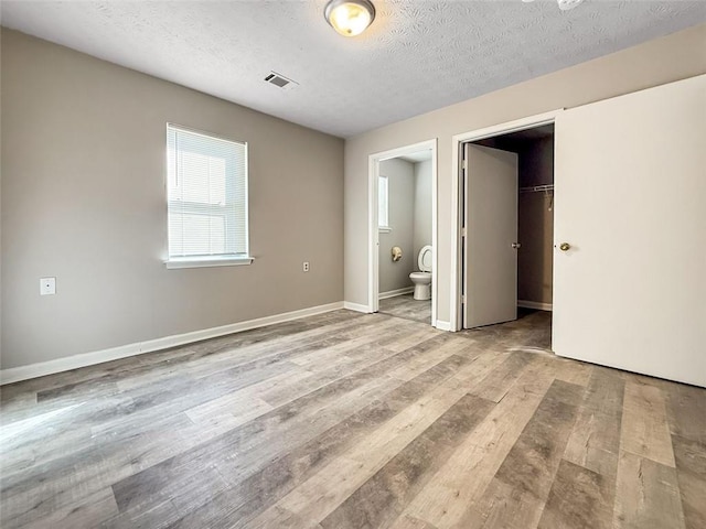 unfurnished bedroom with ensuite bathroom, a spacious closet, a textured ceiling, light wood-type flooring, and a closet