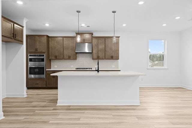 kitchen with double oven, an island with sink, pendant lighting, extractor fan, and ornamental molding