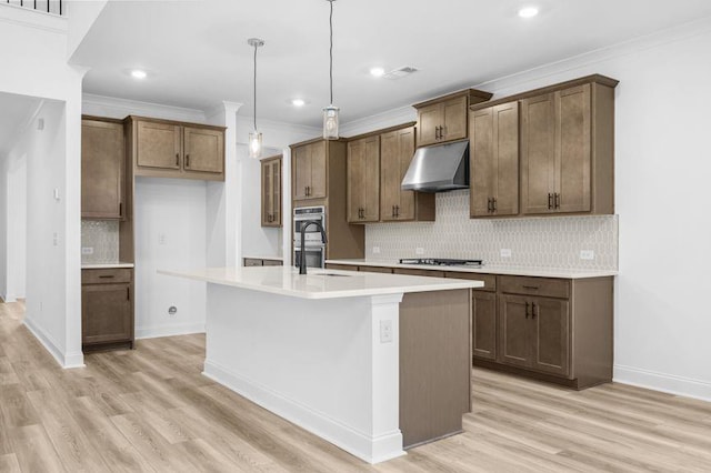 kitchen with appliances with stainless steel finishes, light wood-type flooring, ornamental molding, hanging light fixtures, and an island with sink