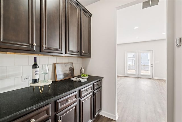 bar with dark brown cabinets, backsplash, dark stone counters, and light hardwood / wood-style flooring