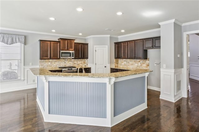 kitchen with appliances with stainless steel finishes, ornamental molding, light stone counters, and an island with sink