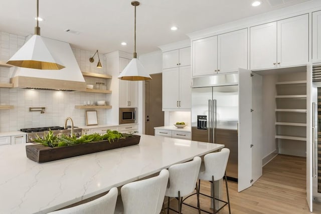 kitchen with white cabinetry, hanging light fixtures, built in appliances, light stone counters, and custom range hood