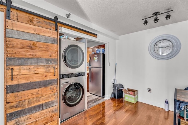 laundry area with a textured ceiling, wood finished floors, stacked washing maching and dryer, a barn door, and laundry area