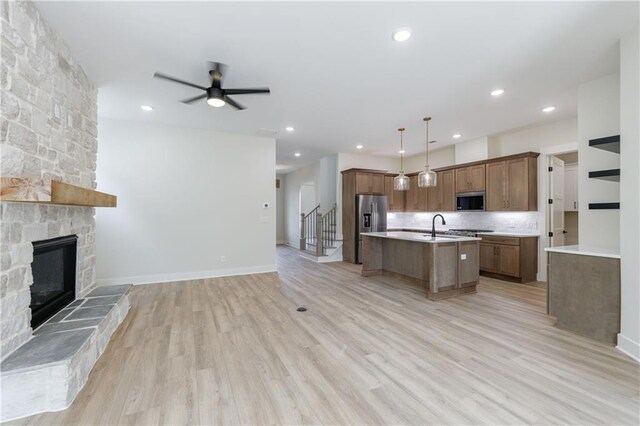 kitchen with stainless steel appliances, a center island with sink, a ceiling fan, a stone fireplace, and open floor plan