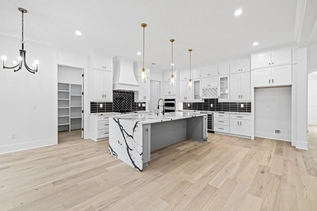 kitchen with white cabinetry, light stone counters, premium range hood, a kitchen island with sink, and light wood-type flooring