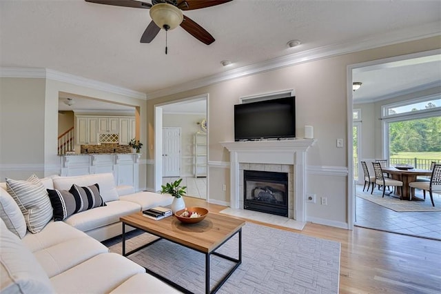 living room featuring ceiling fan, a tiled fireplace, light wood-style floors, ornamental molding, and stairs