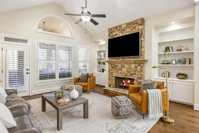 living area featuring built in shelves, dark wood-style floors, ceiling fan, a stone fireplace, and high vaulted ceiling