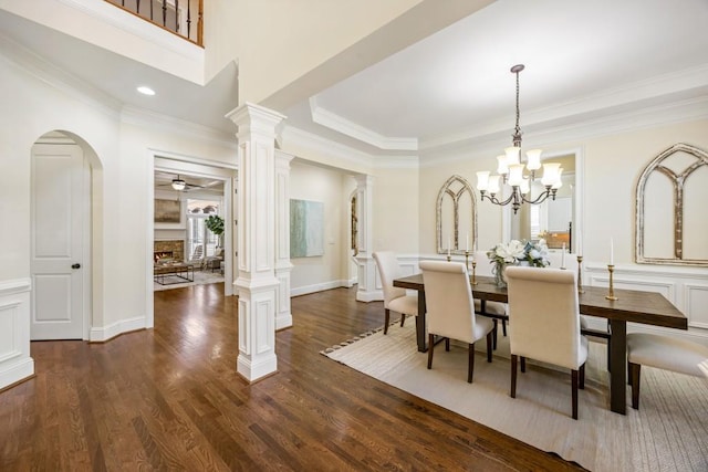 dining area featuring dark wood-type flooring, decorative columns, a lit fireplace, and ceiling fan with notable chandelier