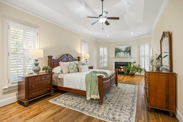 bedroom with baseboards, a glass covered fireplace, wood-type flooring, ornamental molding, and a tray ceiling